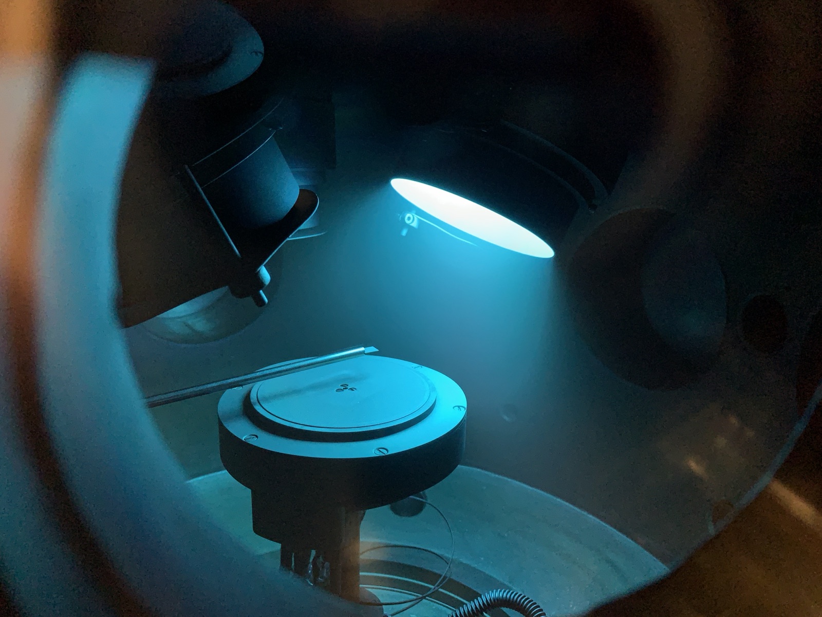 View into a PVD chamber with a sample stage under blue process light.